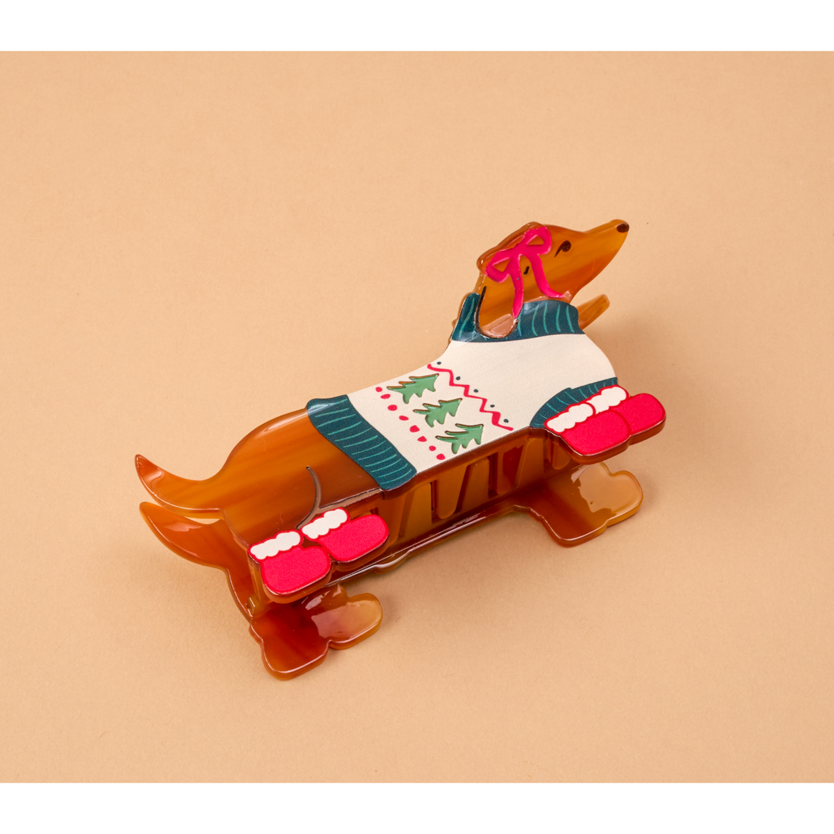 A dachshund figurine wearing a white Christmas sweater with green pine trees and pink boots, set against a solid beige background.