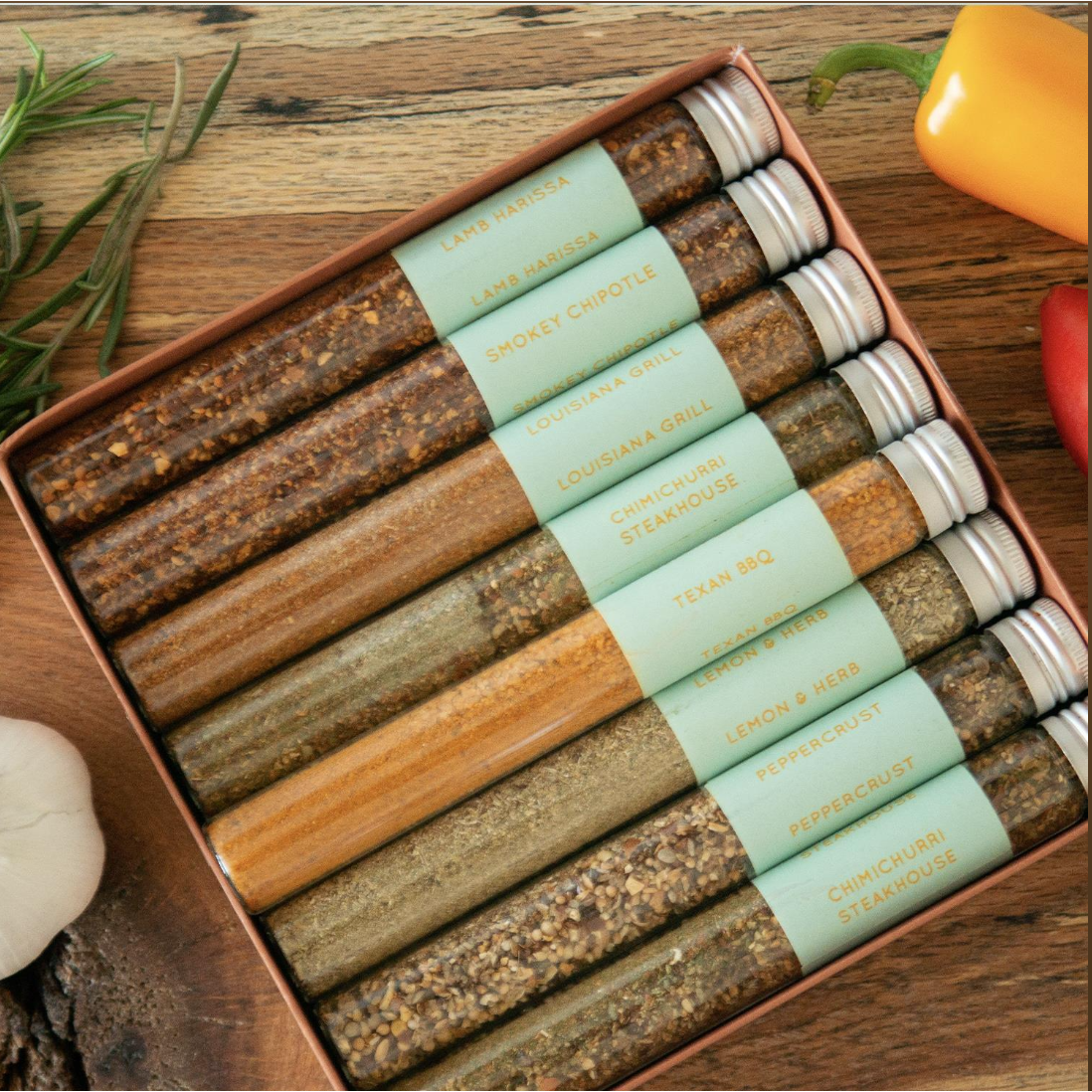 A set of eight spice rubs in clear glass tubes with silver caps and mint green bands, arranged diagonally in a brown box on a rustic wooden table with garlic, rosemary, and peppers.