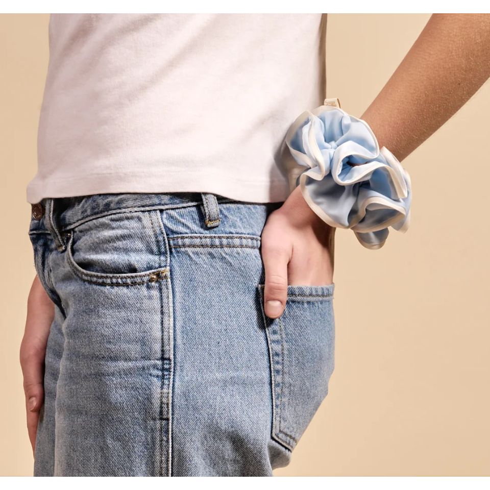 A close-up of a person wearing a light blue satin scrunchie on their wrist, paired with a white t-shirt and light-wash blue jeans, set against a solid beige background.