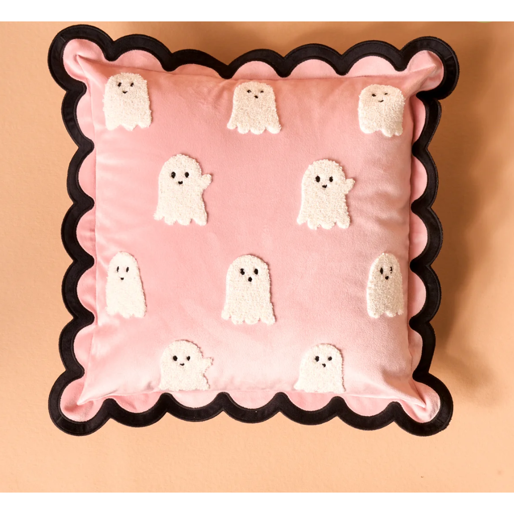 A square pink throw pillow with a black scalloped border and embroidered white ghost motifs, photographed against a warm peach background.