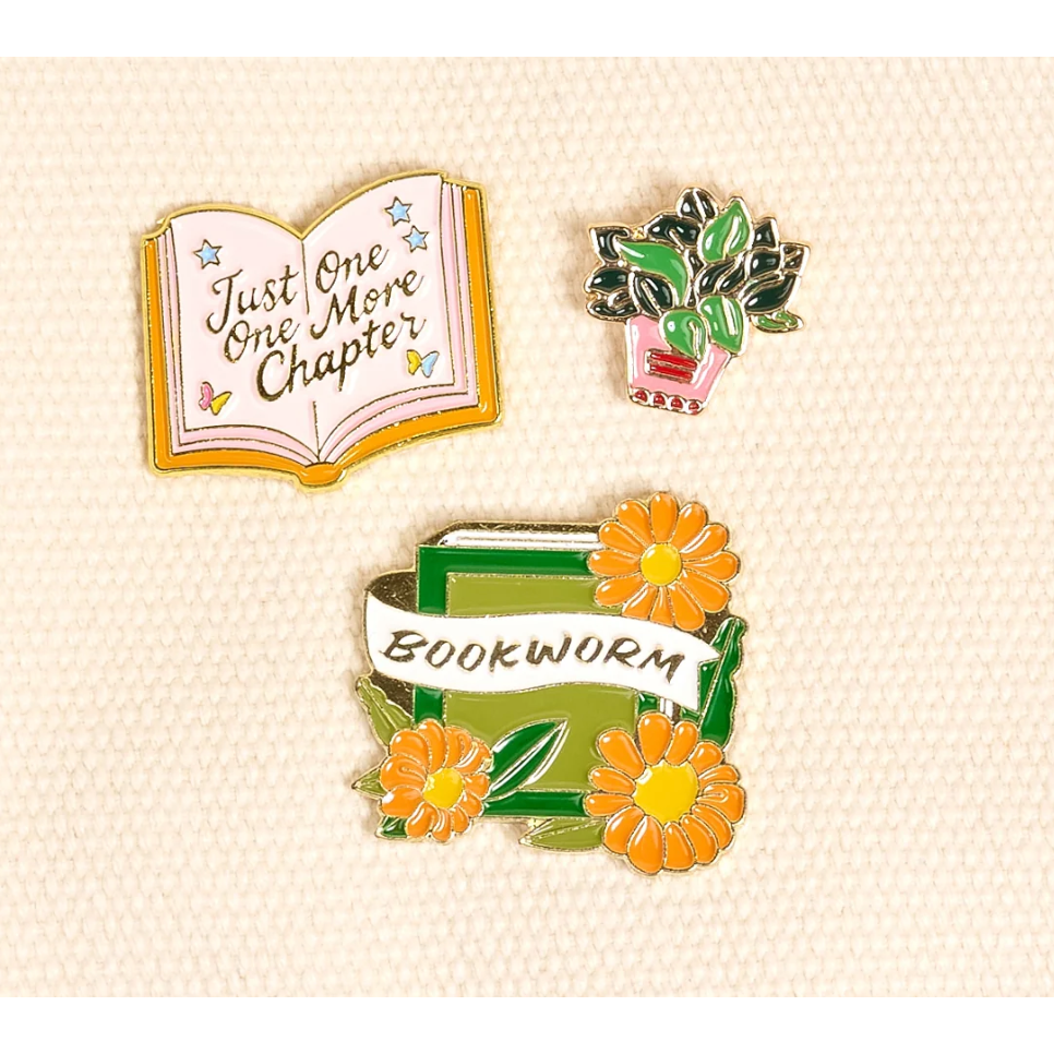 Three small enamel pins displayed on a light cream textured background: an open book pin reading "Just One More Chapter," a potted plant pin, and a bookworm pin with orange flowers and the word "BOOKWORM.".