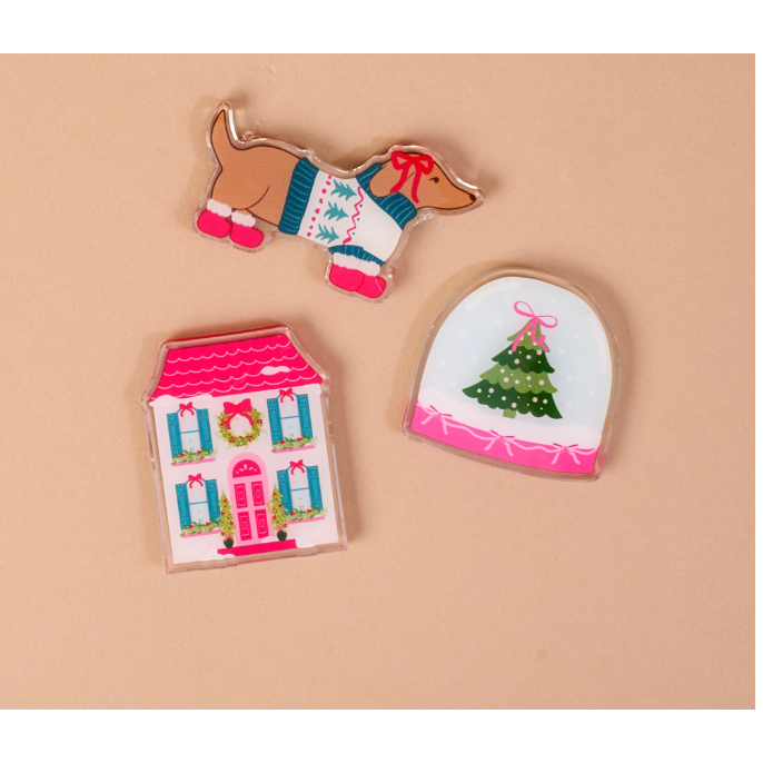 Three Christmas-themed enamel pins on a solid beige background: a dachshund wearing a sweater and pink boots, a pink house with blue shutters and a wreath, and a white arched shape featuring a green Christmas tree with a pink bow.