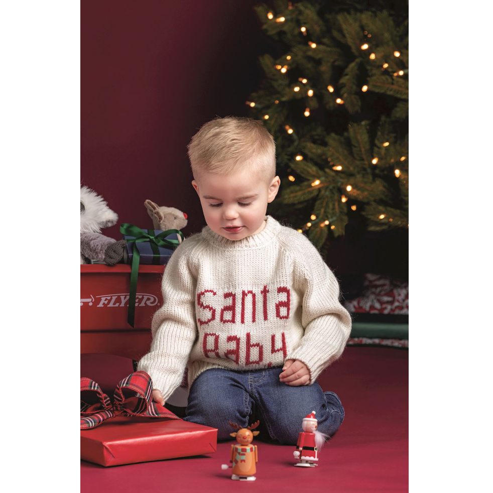 Child wearing a 'Santa Baby' sweater sitting on the floor with Christmas decorations.