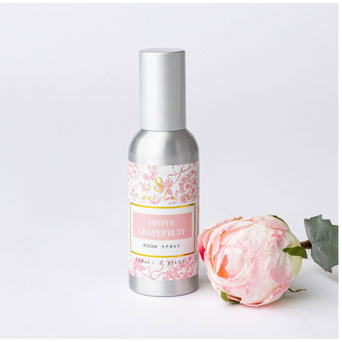 Room spray bottle with floral label next to a pink flower on a white background