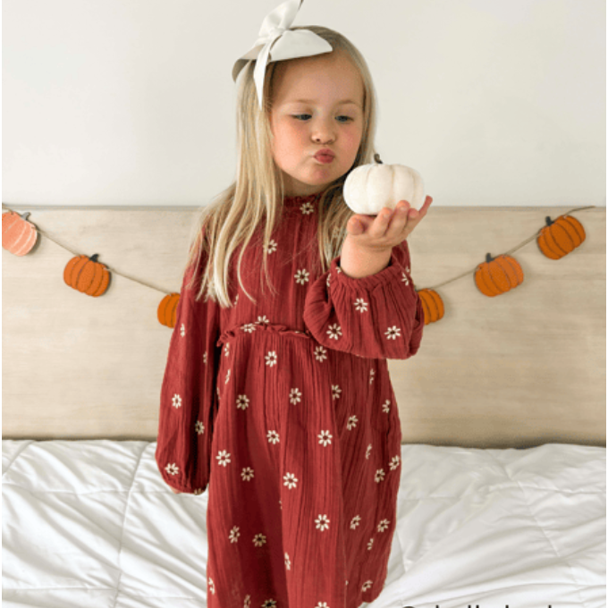 Child in a red dress holding a white pumpkin with a pumpkin-themed headband and decorations in the background.