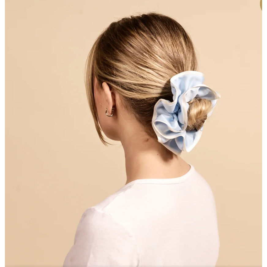 A woman shown from the back wearing a light blue satin scrunchie in her hair, paired with a white top against a solid beige background.