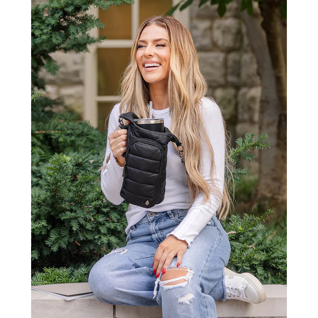 Woman holding a black jacket outdoors with greenery and a stone wall in the background