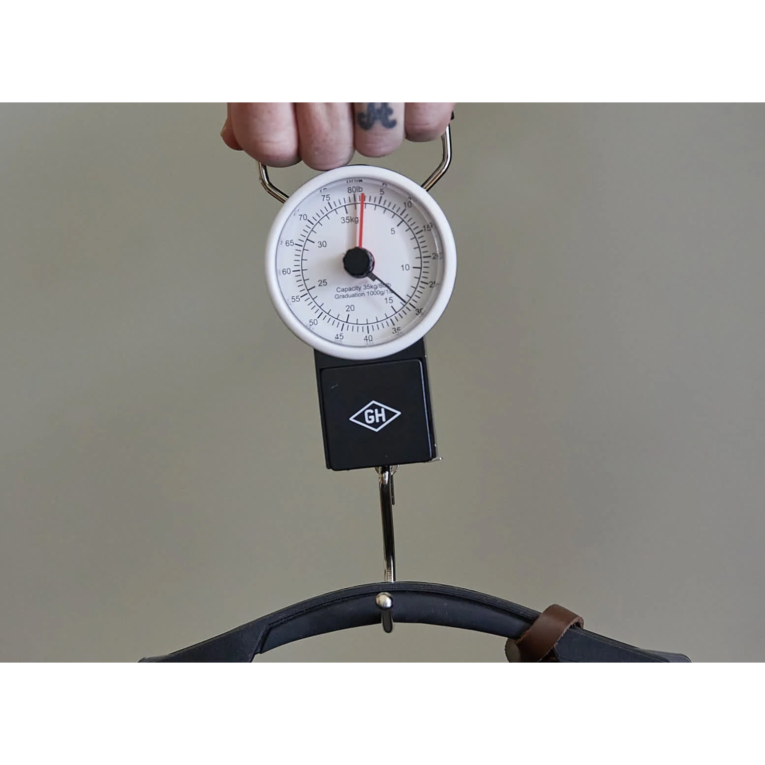 A hanging analog scale with a white face, black housing, and metal hook, being held by a hand against a plain beige background.