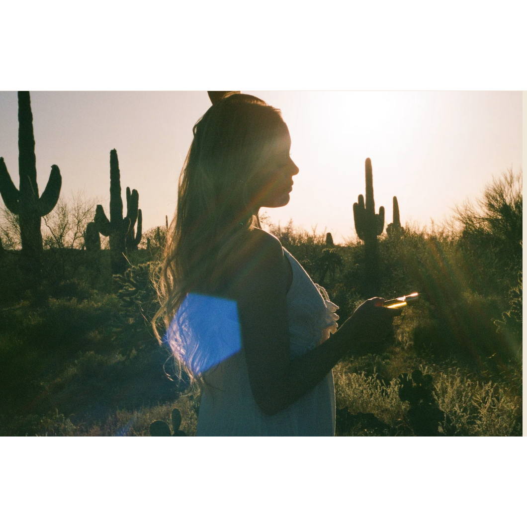 A woman in a white sleeveless dress stands in profile in a desert landscape with saguaro cacti, backlit by the setting sun and holding a phone.
