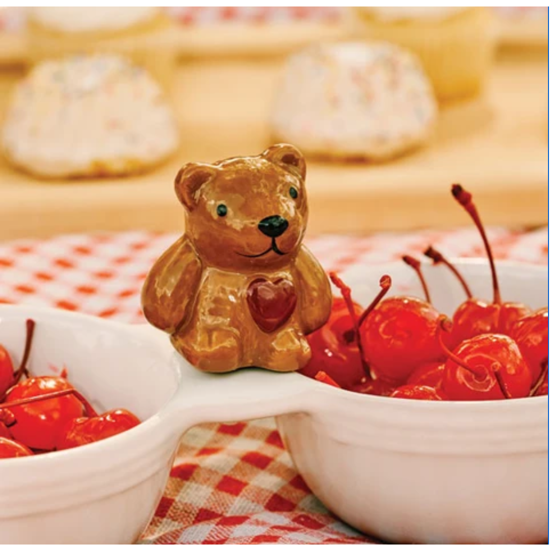 A small brown ceramic bear figurine with a red heart on its chest, placed on the handle of a white two-bowl serving dish filled with red cherries, with a blurred background of cupcakes and a red-and-white checkered cloth.