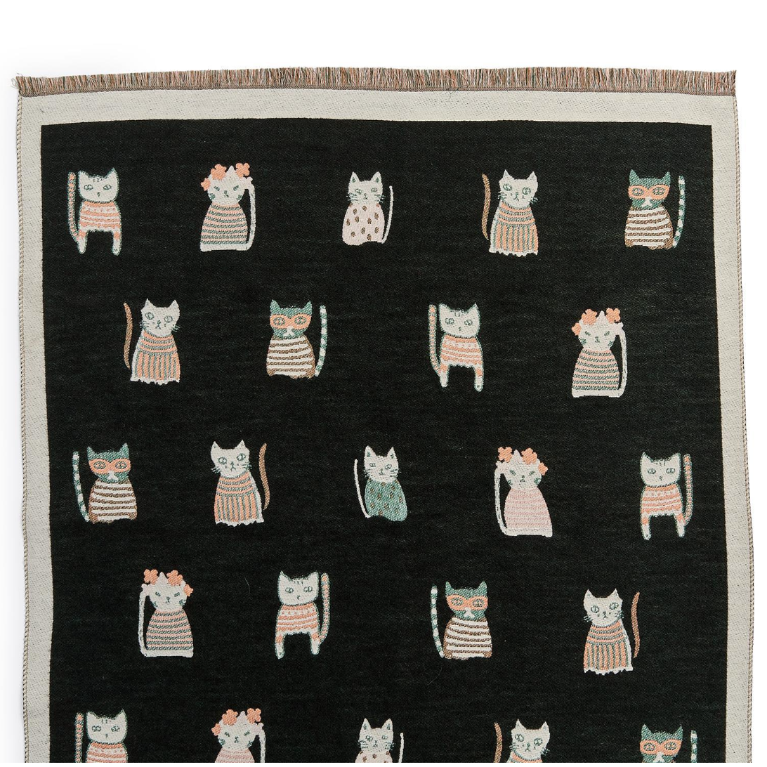 A black woven throw blanket with a fringed top edge, featuring a repeating pattern of stylized cats in pink, beige, and light blue-gray outfits.