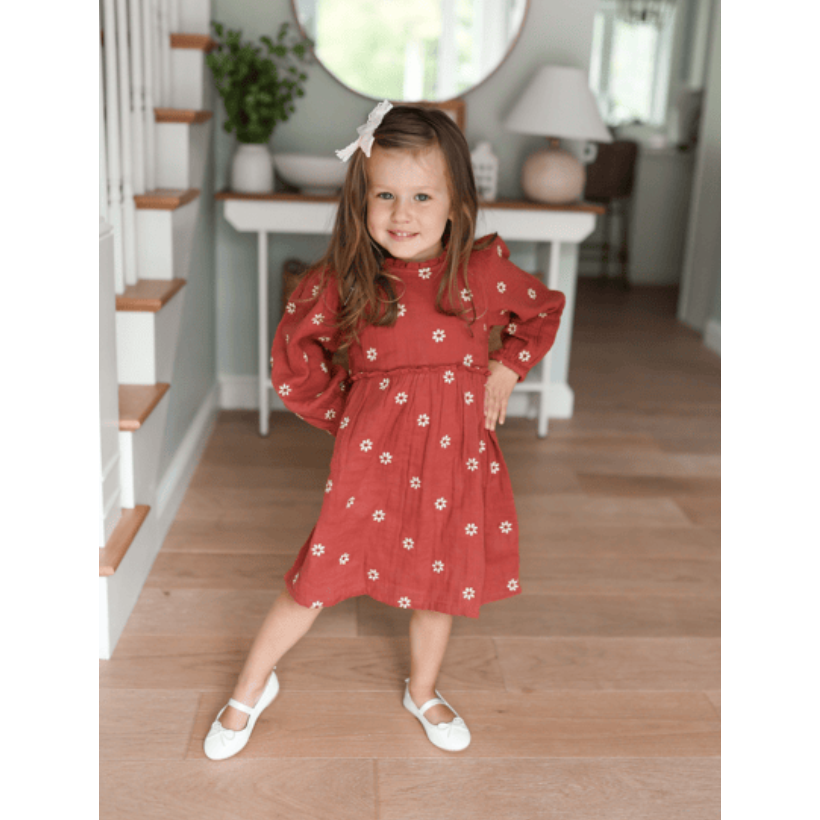 Young girl in a red dress standing on a wooden floor with a staircase and table in the background.