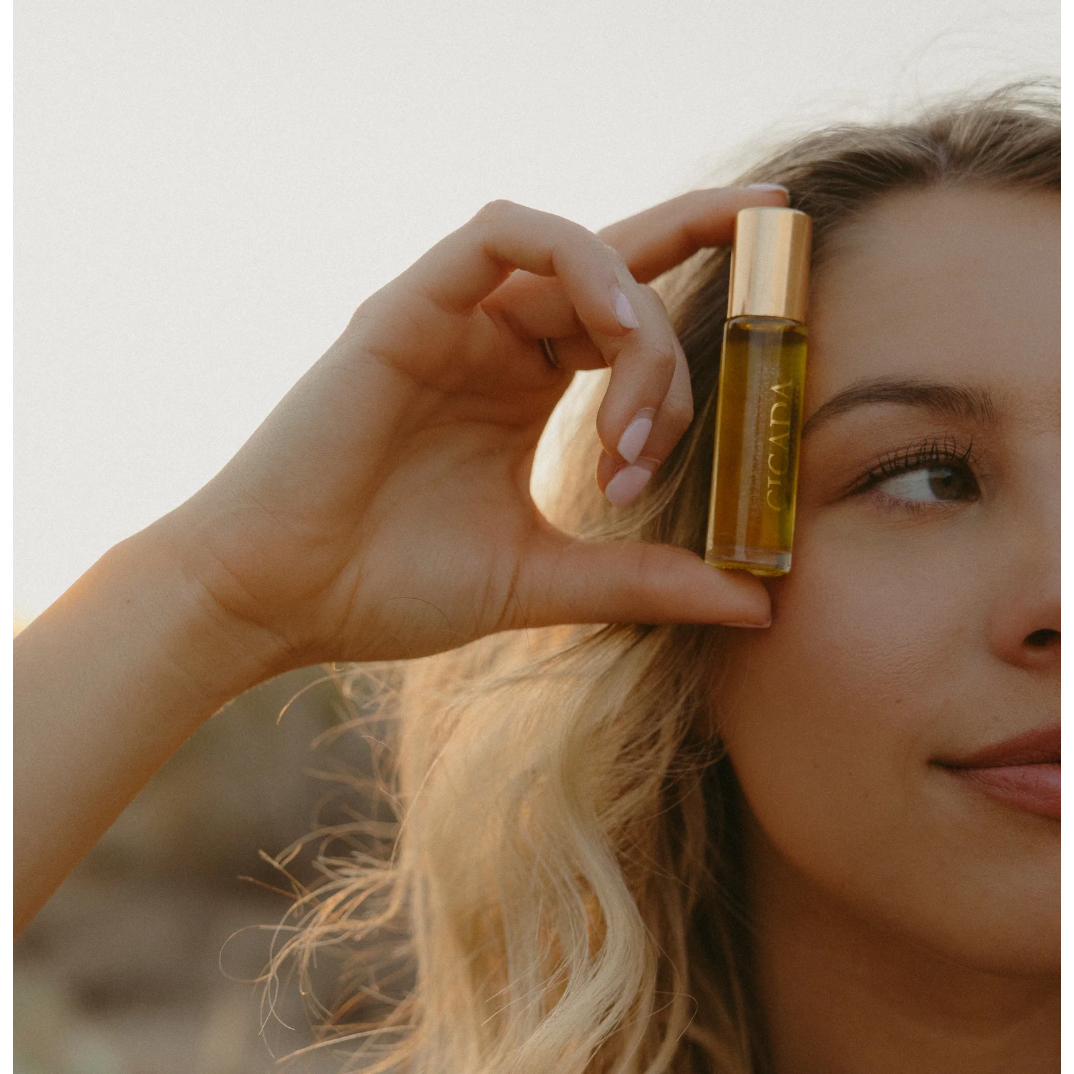A close-up lifestyle shot of a woman holding a small glass rollerball bottle with a gold cap next to her face. The bottle contains a golden-yellow liquid and has the brand name 'VIVID' printed vertically on it.