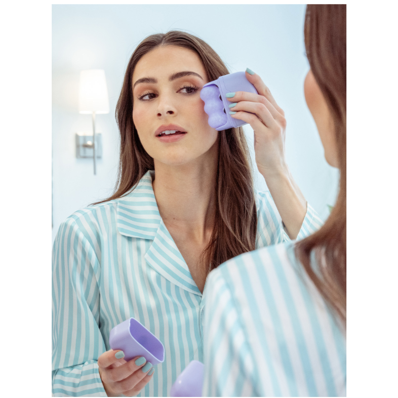  woman in a bathroom wearing a light blue and white striped pajama shirt uses a lavender silicone face mask applicator while looking into a mirror.