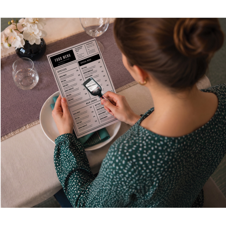 A woman viewed from behind at a restaurant table, holding a printed menu and a small black tablet over it. She wears a dark green long-sleeve top with a small white pattern.