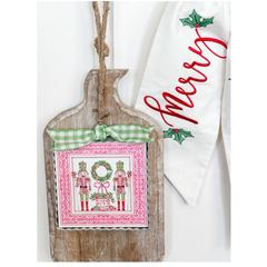 Decorative Christmas card with nutcrackers, wreath, and candy canes on a wooden board with a green ribbon, next to a towel with 'Merry' text.