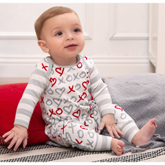 Baby wearing a white onesie with red and gray patterns, sitting on a bed.