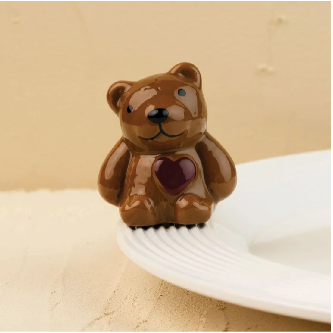A small glossy brown bear-shaped object with a dark red heart on its chest, shown sitting on a white plate against a warm beige background.