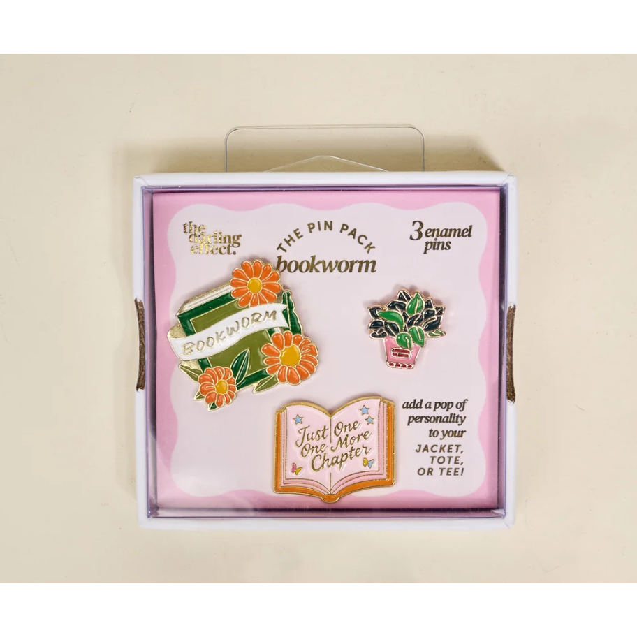 A boxed enamel pin set on a light beige background. The package is pink and white with a clear plastic hanger at the top and shows three book-themed pins: a bookworm with orange flowers, a potted plant, and an open book with the phrase 'Just One More Chapter'.