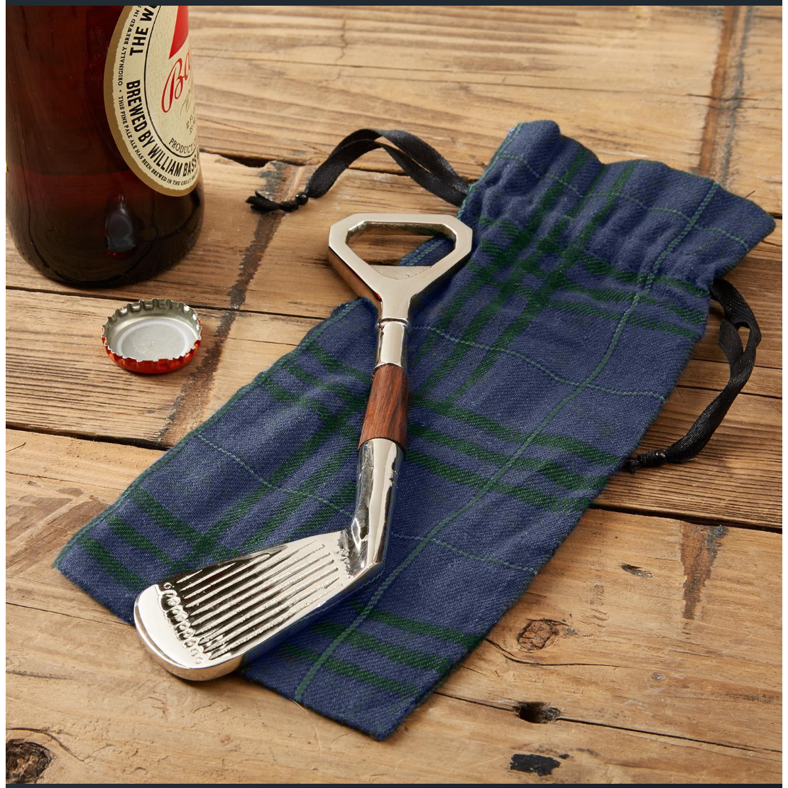 A metal bottle opener shaped like a golf club lies on a blue plaid fabric pouch with black drawstrings, placed on a rustic wooden tabletop. Part of a brown bottle and a red cap are visible at the upper left.