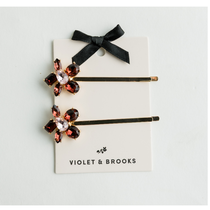 Two black bobby pins with flower-shaped crystal embellishments in dark pink and gold, attached to a white display card with a black ribbon bow and the brand name VIOLET & BROOKS.