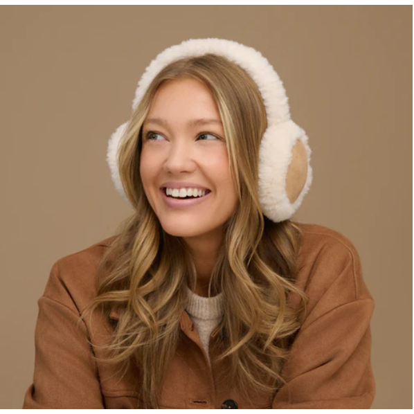 A woman smiling while wearing fluffy white earmuffs with tan ear pads, photographed against a plain warm beige background.
