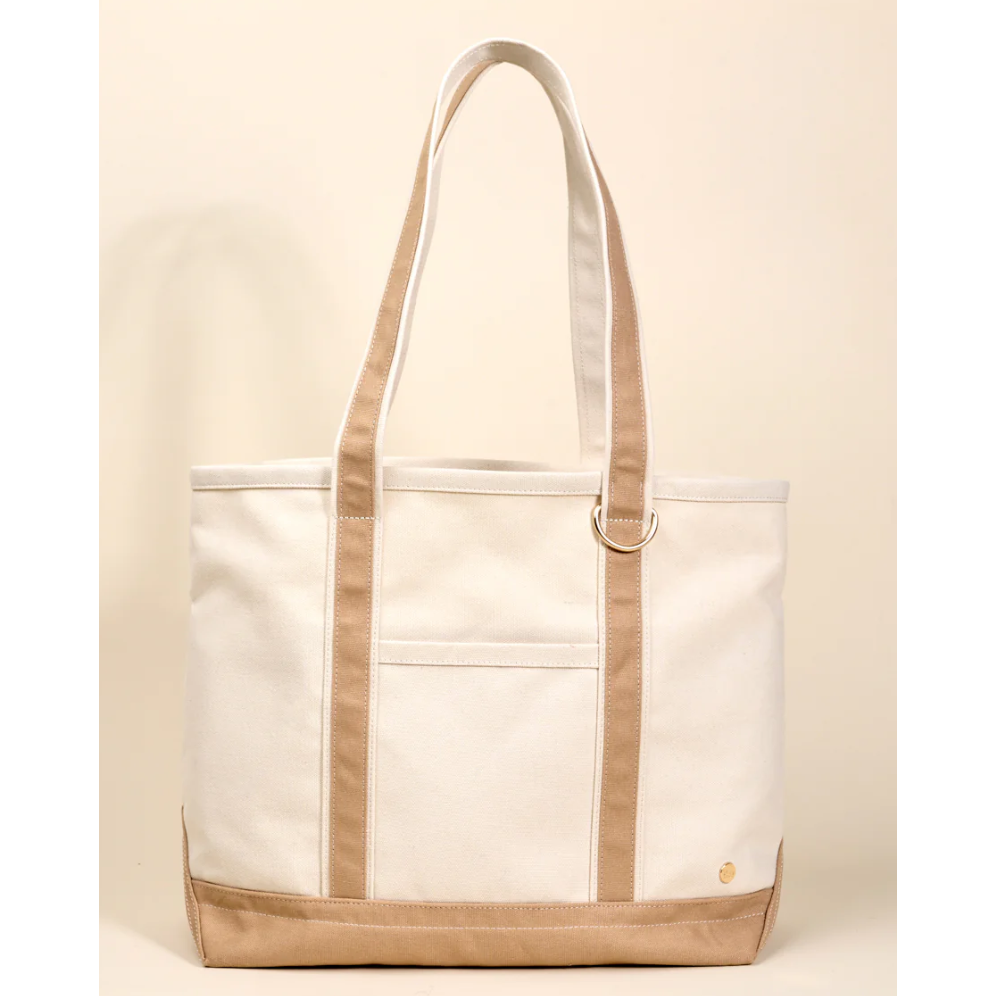 A canvas tote bag with off-white body panels and tan trim, including the handles, side straps, and bottom base. It features a front slip pocket and a small gold logo on the lower right corner, set against a solid cream background.