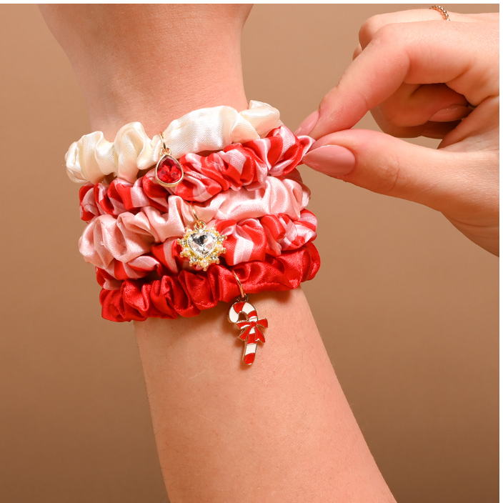 A close-up of a person's wrist wearing a stack of satin scrunchie-style bracelets in red, pink, and white, with decorative charms including a small gold heart and a candy cane.