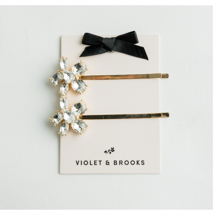 Two gold-tone hair pins with floral crystal embellishments and a black ribbon bow, displayed on a white card printed with the brand name 'VIOLET & BROOKS'.