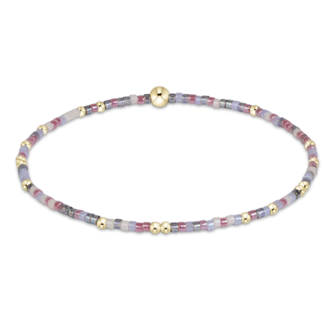 A delicate beaded bracelet with small lavender, pink, and gray stone beads interspersed with polished gold spacer beads and a large round gold bead, shown on a plain white background.