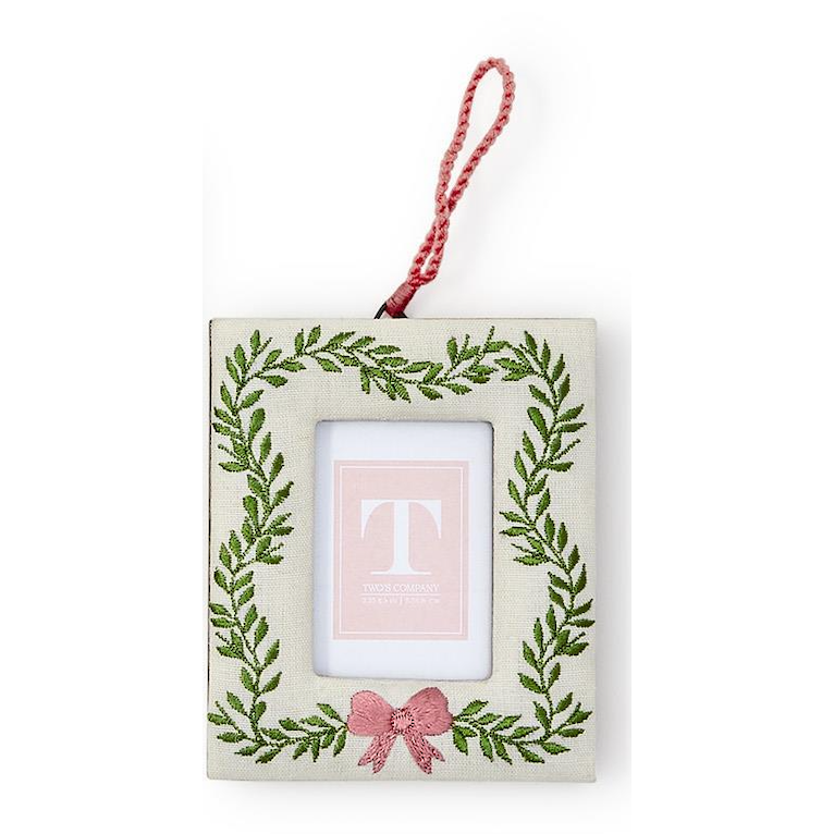 A square fabric hanging ornament with a red hanging loop, featuring an embroidered green leaf wreath border, a pink bow at the bottom, and a central photo slot with a pink insert showing a monogram letter 'T'.