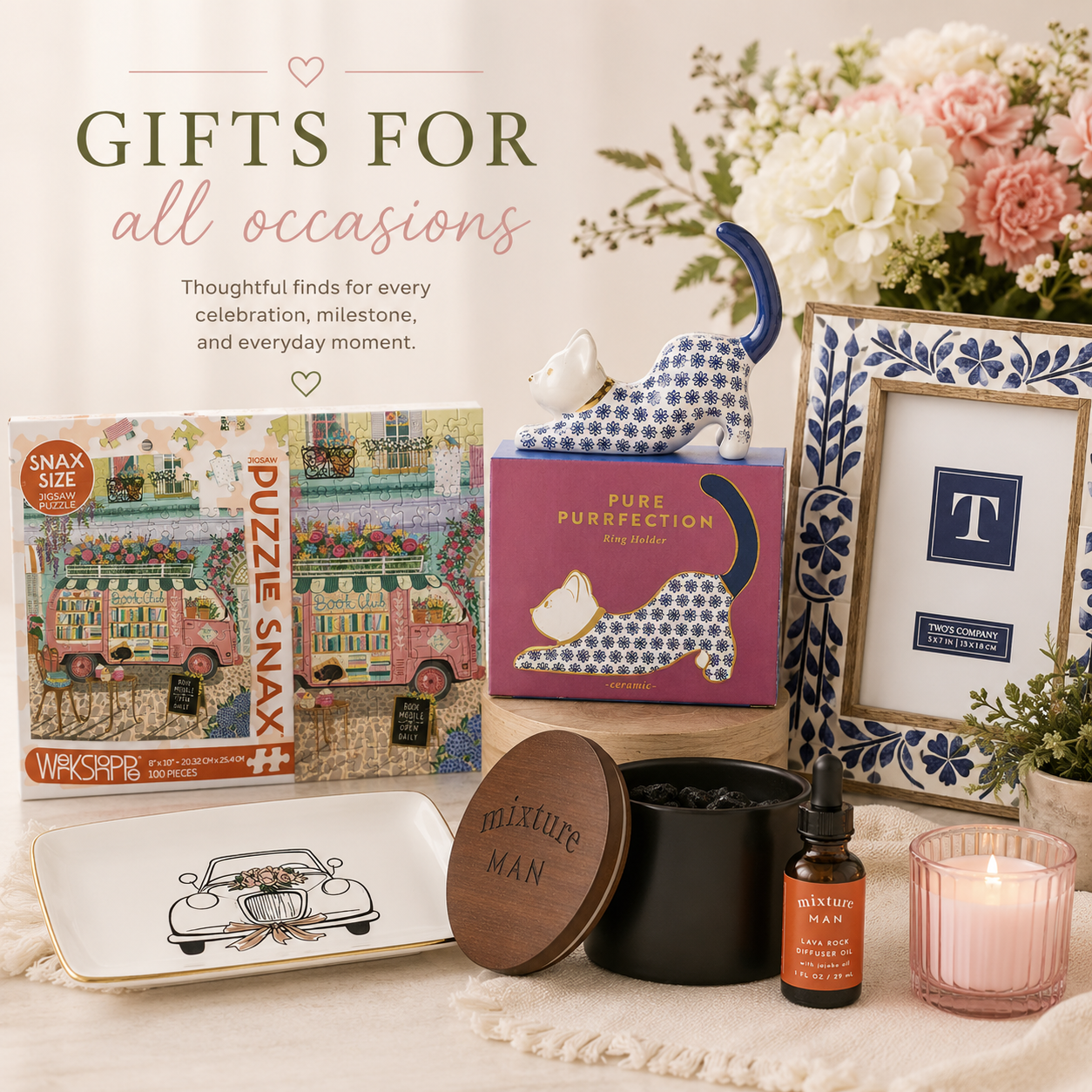A curated gift set on a cream fabric surface with floral decor. Visible items include a Wacko! Puzzle Snax jigsaw puzzle box, a pink cat figurine in a box labeled Pure Purification, a framed monogram letter T, a small bottle labeled Bluntary Nail, a pink candle, a black container with a round wooden lid reading Mixteer Man, and a white tray with a car illustration.