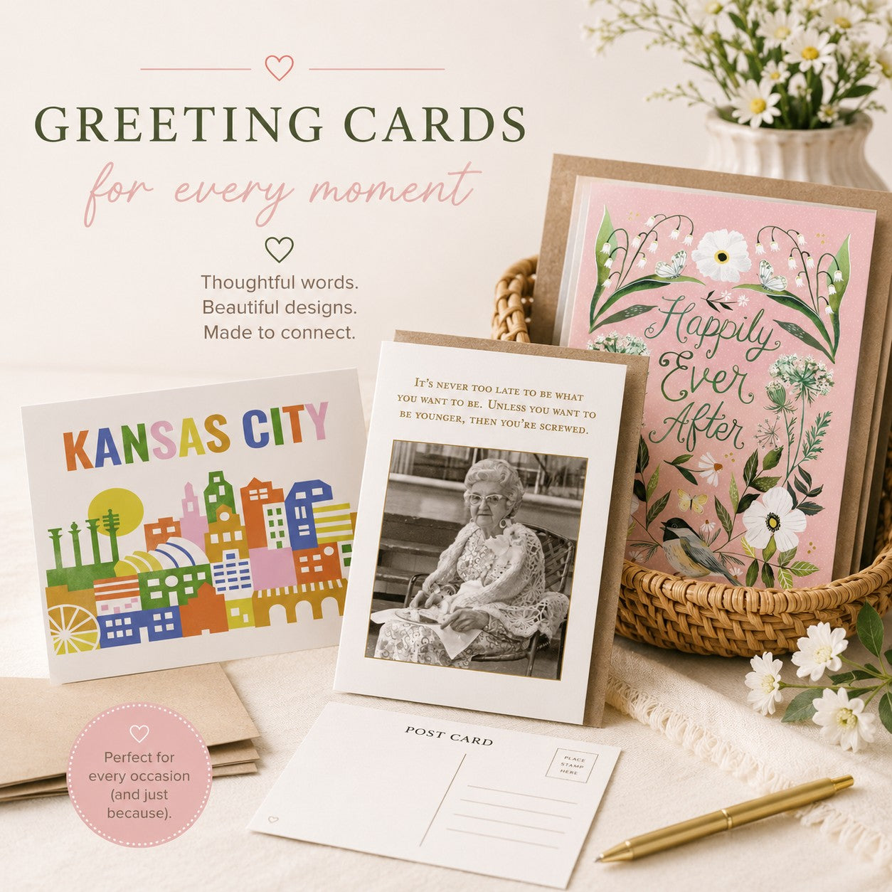 A styled product display of greeting cards on a soft cream fabric background. Visible items include a pink floral card reading 'Happily Ever After,' a colorful 'KANSAS CITY' cityscape card, a postcard with a black-and-white portrait and quote, a postcard back, a pen, a basket, and decorative flowers.