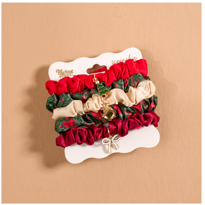 A set of Christmas-themed fabric hair scrunchies on a white display card. The scrunchies include solid red, green with a red pattern, solid beige, and deep burgundy velvet, with a small gold bell charm and a silver bow charm.