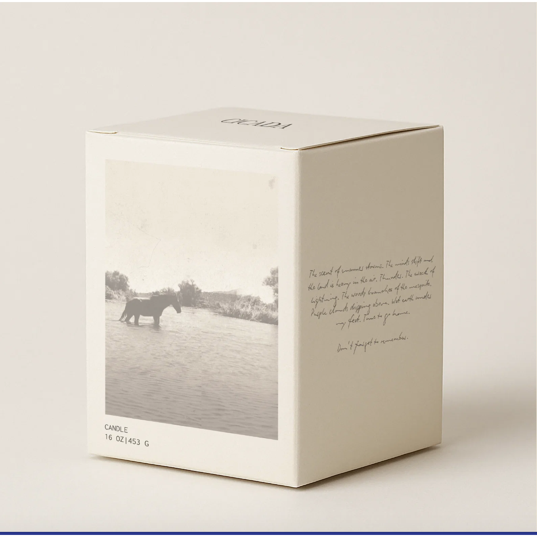A cream-colored square candle box with a printed vintage-style photograph of a horse in water on the front and elegant cursive text on the side, photographed on a neutral light beige background.
