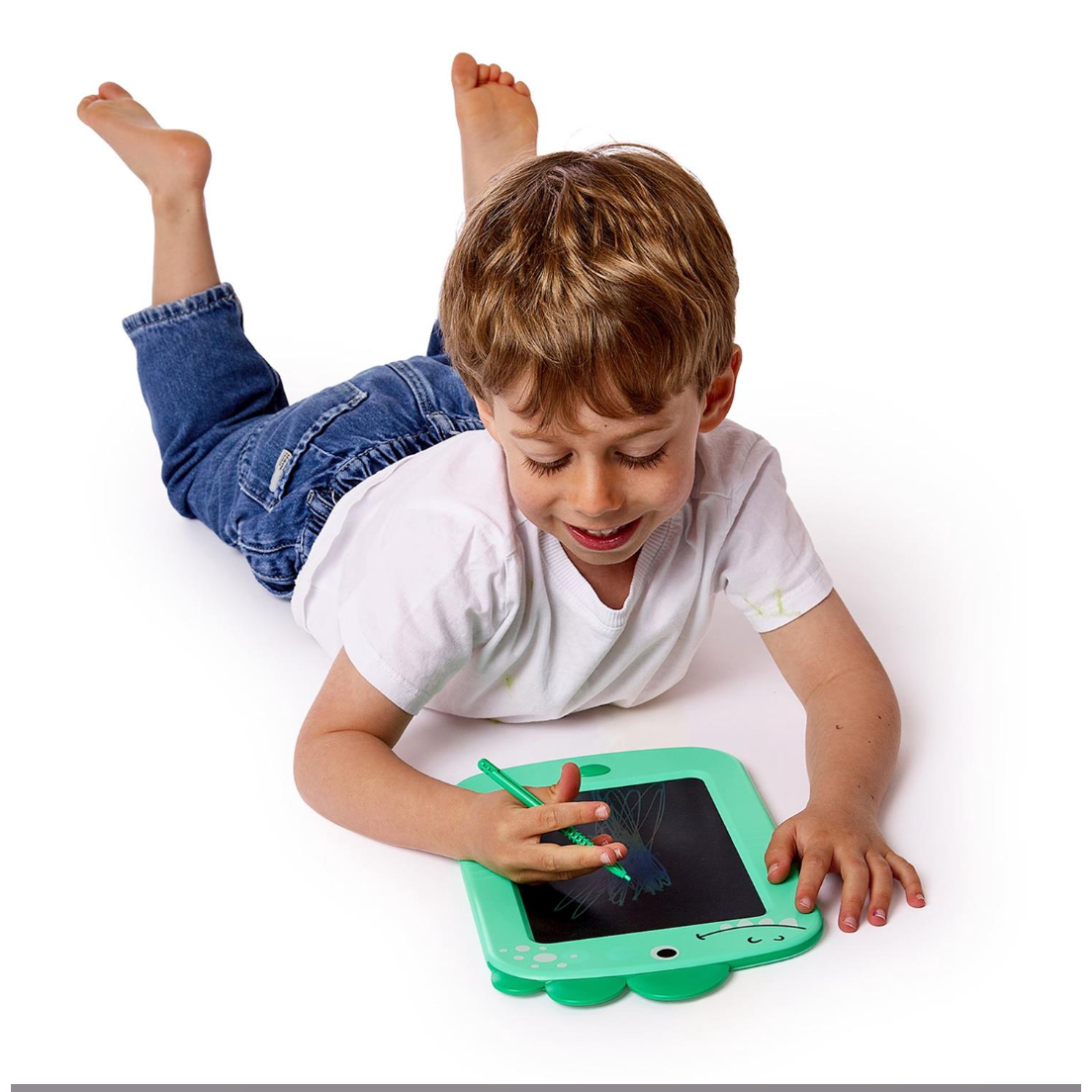 A young boy with blond hair lies on his stomach on a white floor, using a mint green tablet-style writing pad with a matching stylus. He is wearing a white t-shirt and blue jeans.