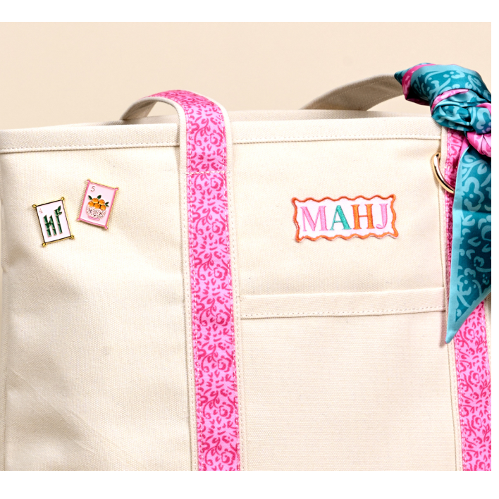 A cream canvas tote bag with bright pink patterned trim and handles, featuring a colorful embroidered 'MAHJ' patch on the front pocket and two small decorative pins on the left side. A teal patterned scarf is tied to the handle.