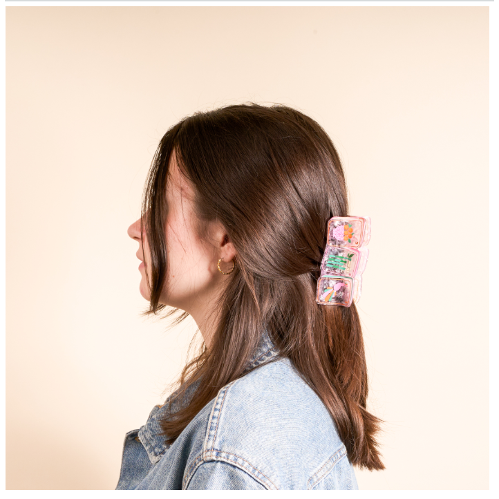A woman in profile wearing a light blue denim jacket and a pink floral hair claw clip, shown against a solid beige background.