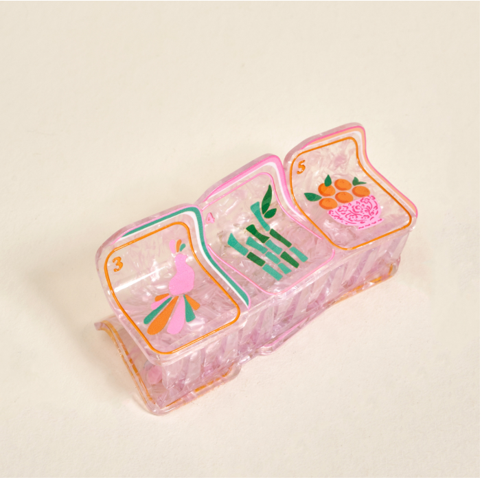 A translucent pink plastic hair claw clip with three compartments decorated with colorful illustrations of a pink flamingo, green bamboo, and orange fruits, set against a plain beige background.