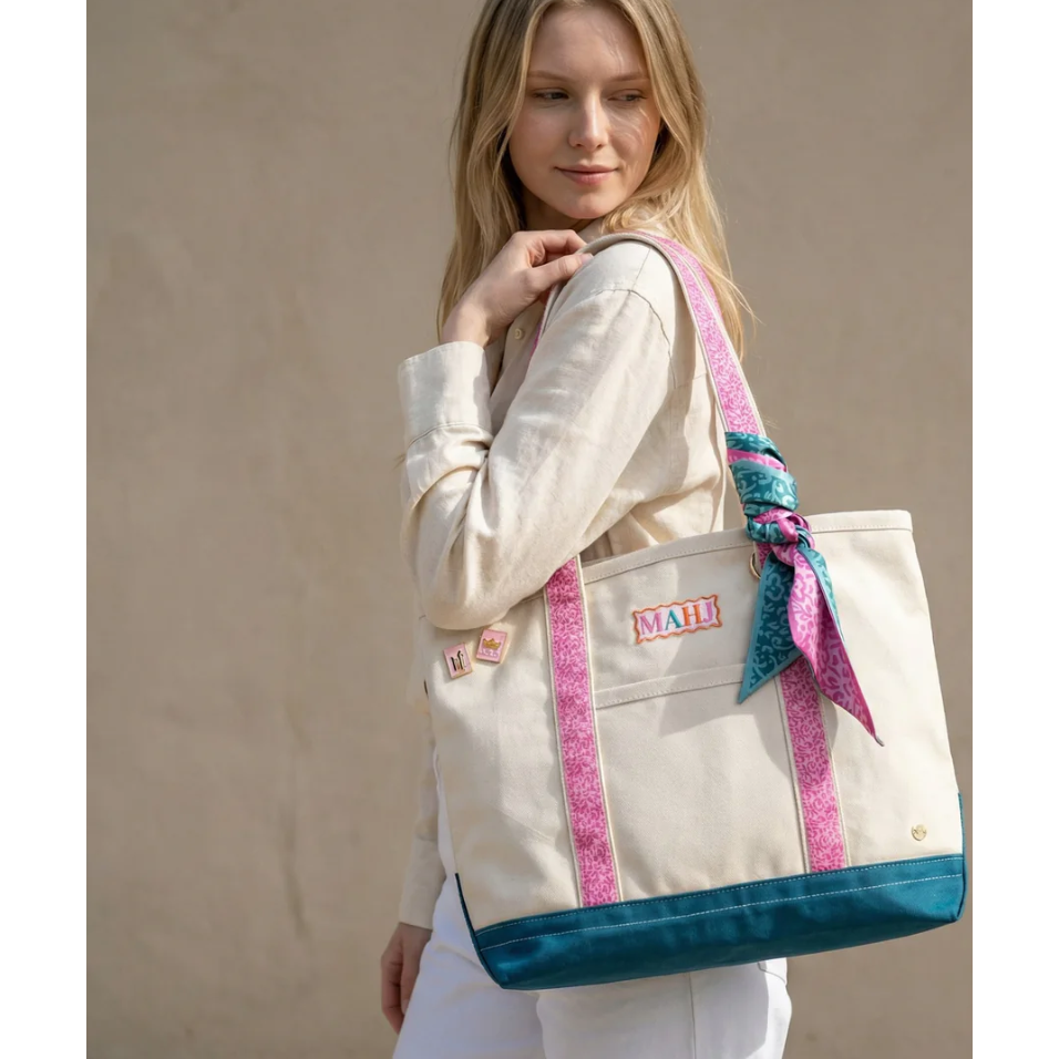 A woman wearing a cream linen shirt and white pants carries a large cream canvas tote bag with a teal bottom, pink patterned straps, and a matching pink and teal scarf tied to the strap. The tote has a front pocket with a colorful 'MADE IN USA' patch and a small brand patch that reads 'MADE'.
