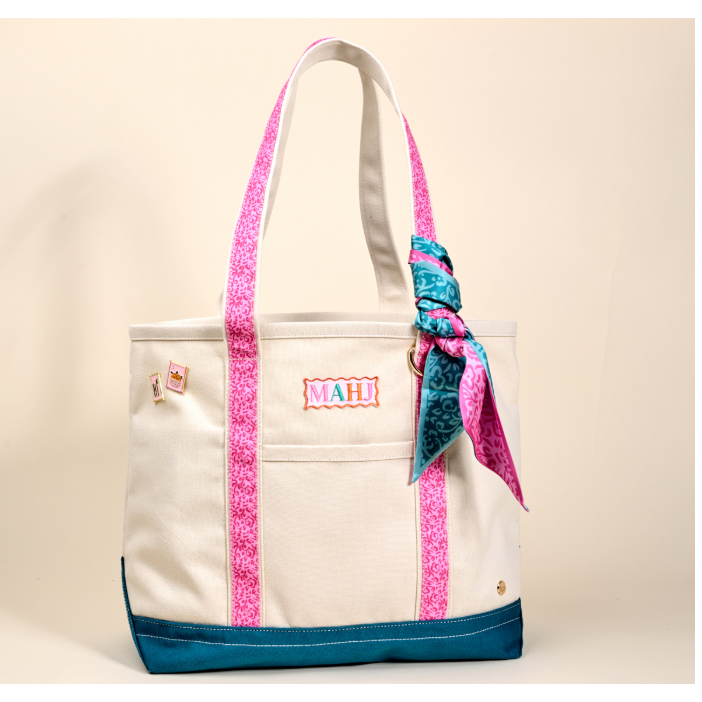 A cream canvas tote bag with bright pink patterned handles and side trim, a teal bottom panel, and a matching pink and teal ribbon tied in a bow on the side. The front pocket has a colorful 'MAHI' patch.
