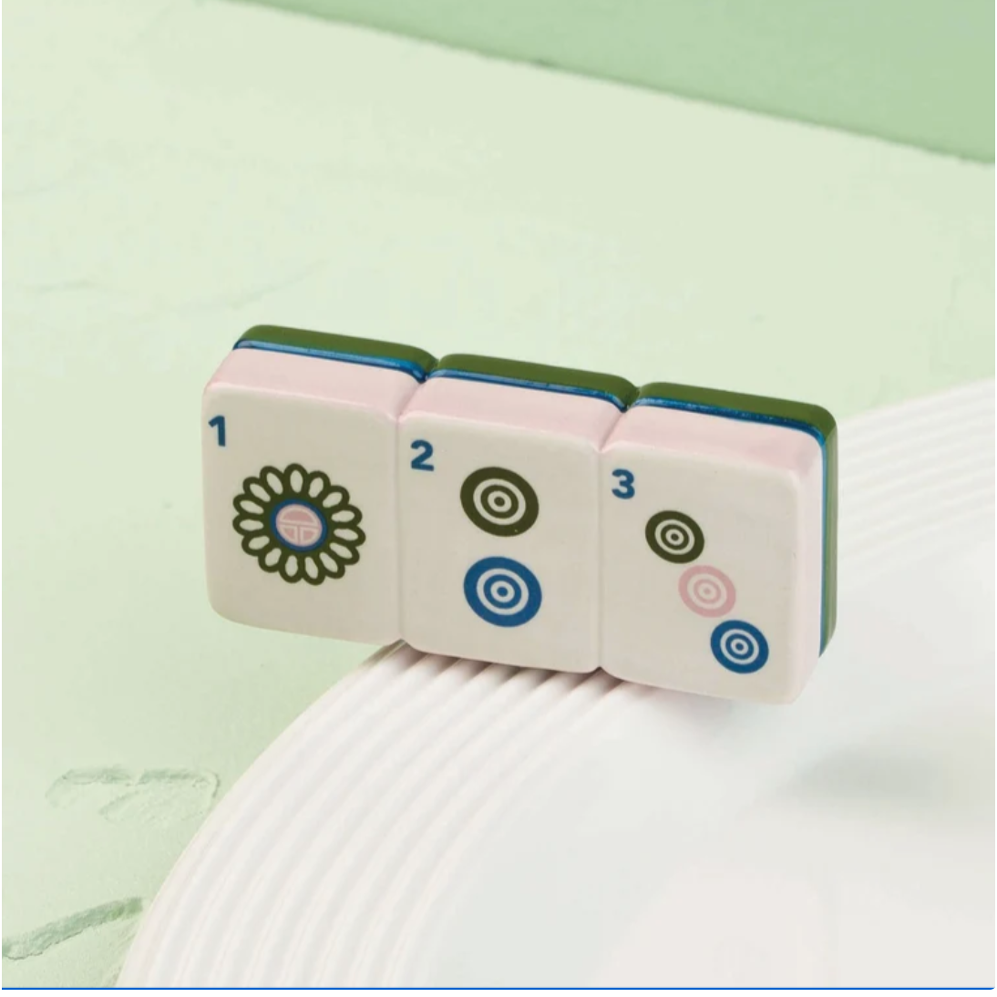 Three miniature Mahjong-style tiles with printed numbers 1, 2, and 3 rest on a white ribbed surface against a pale mint green background.
