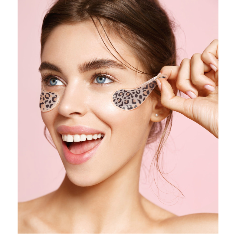 A woman with leopard-print under-eye patches on her eyes, holding one patch with her hand against a solid pink background.