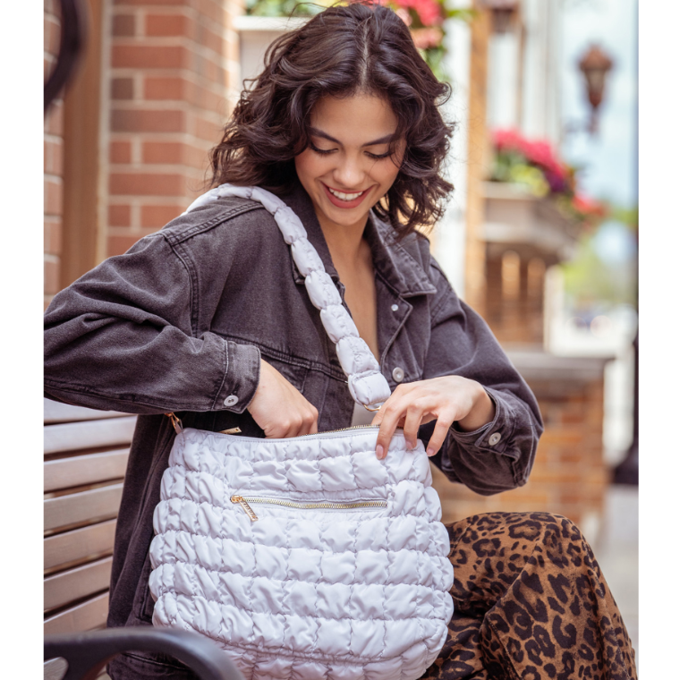 A woman in a dark denim jacket and leopard-print pants smiles while sitting on a bench, showing the spacious zippered pocket of a light gray quilted shoulder bag.