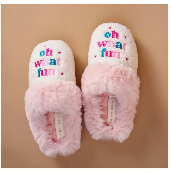 A pair of fluffy slip-on slippers in white and light pink, featuring the phrase 'oh what fun' embroidered in colorful pink, blue, and red letters on the top. The slippers are displayed on a solid tan background.