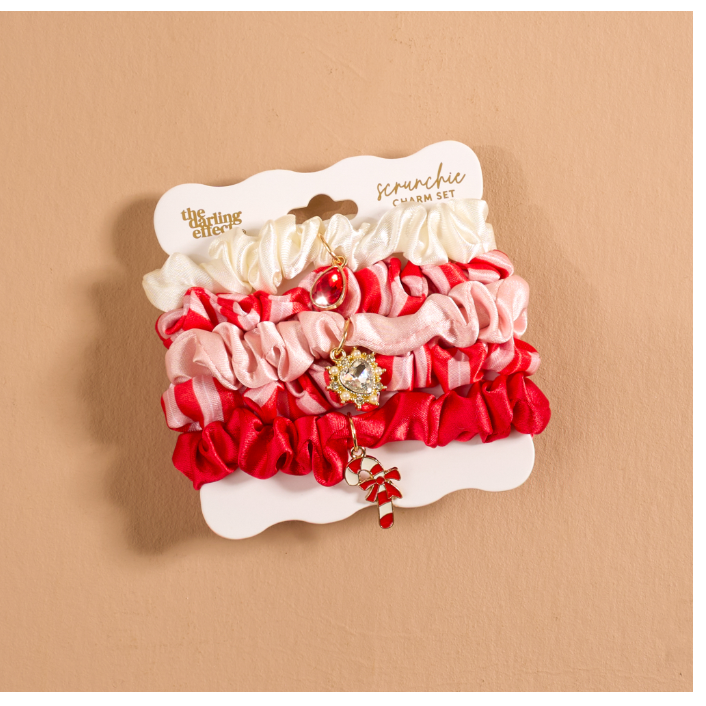 A set of four satin ruffled charm headbands in white, light pink, medium pink, and red, packaged on a white display card with gold lettering. The headbands feature decorative crystal and bead charms, and are displayed against a solid tan background.