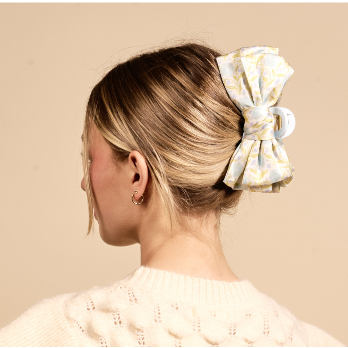 A woman shown from the back wearing a large fabric hair bow hair clip in a pale yellow and white pattern, holding her hair in a low bun. She is wearing a cream-colored knit sweater against a solid beige background.