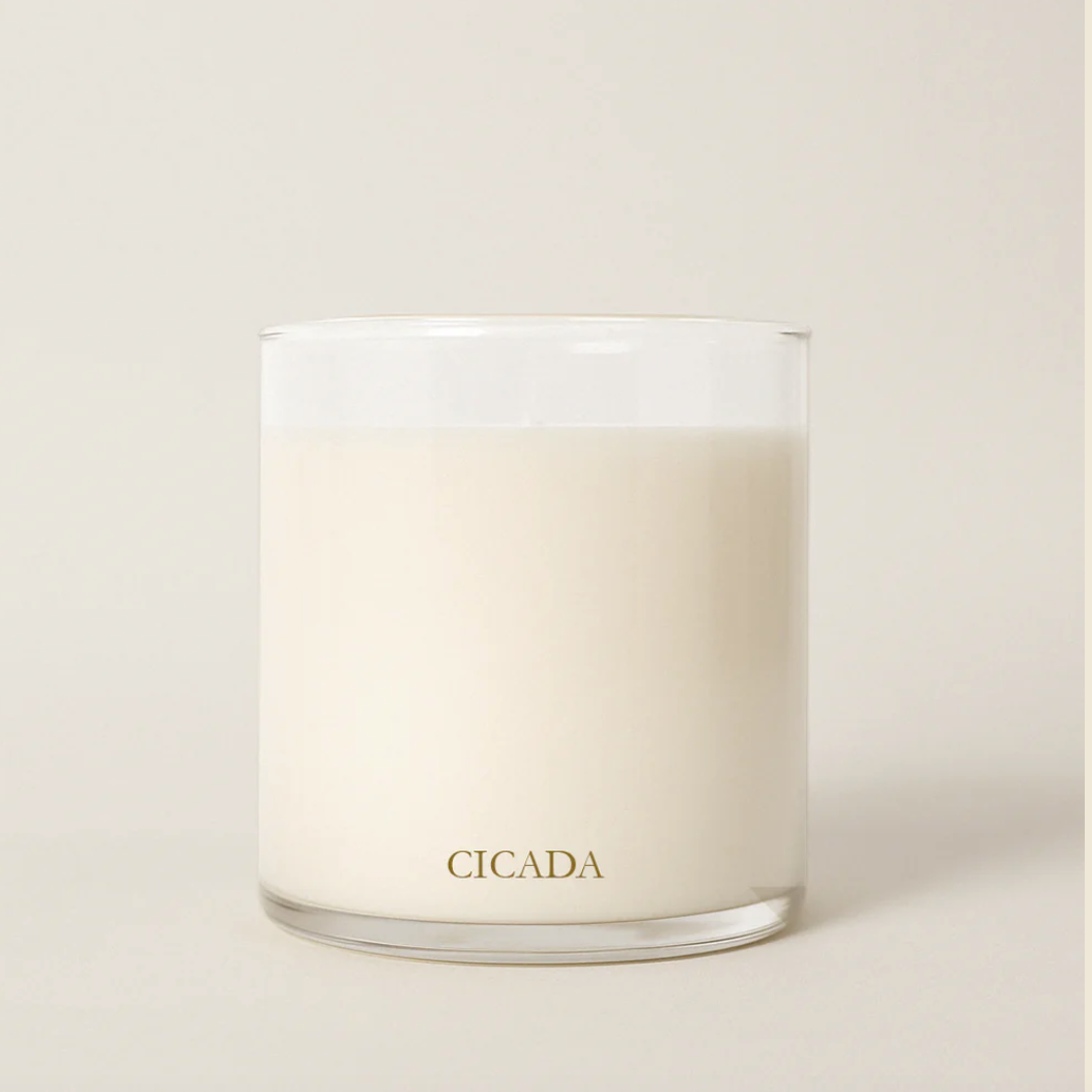 A clear glass jar candle filled with creamy off-white wax, featuring the brand name 'CICADA' printed in gold at the bottom, set against a solid light beige background.