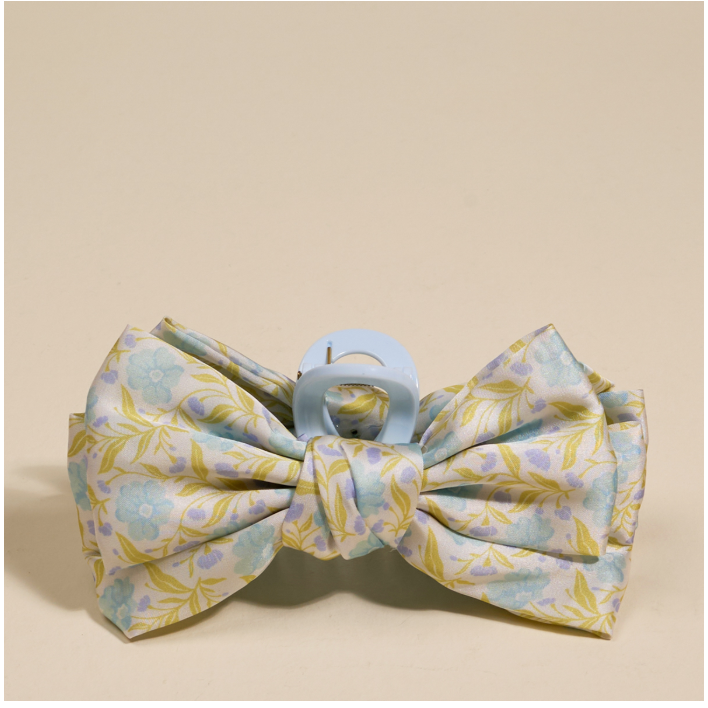 


A fabric hair bow with a light blue and yellow floral pattern, attached to a light blue plastic claw clip, shown on a solid beige background.