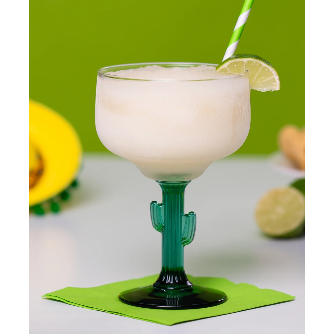 Cactus-shaped glass with a creamy drink, lime wedge, and straw on a green background
